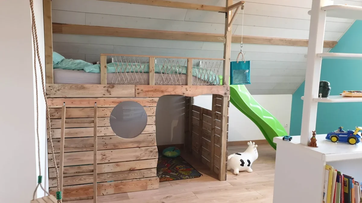 Lit cabane surélevé sur mesure en bois brut finalisé dans une chambre d'enfant mansardée, avec hublot, filets de sécurité et rangements intégrés, par Garcia Rénovation à Angers.